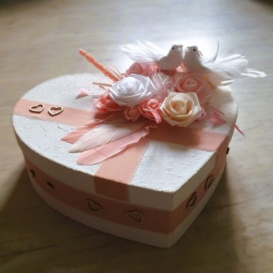 Boîte en forme de cœur décorée de fleurs et de rubans, avec deux petites figurines.