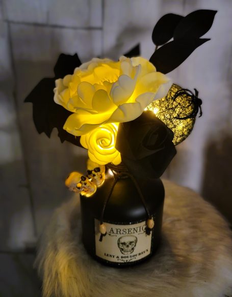 Vase noir décoré de fleurs blanches et jaunes, avec des feuilles sombres et des lumières.