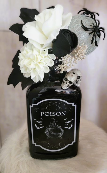 Bouteille noire décorée de fleurs blanches, araignées et une tête de mort.