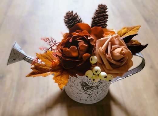 Bouquet de fleurs artificielles dans un arrosoir en métal, avec des feuilles et des cônes.