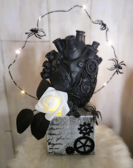 Cœur noir décoré de mécanismes, fleurs blanches et araignées, avec guirlande lumineuse.